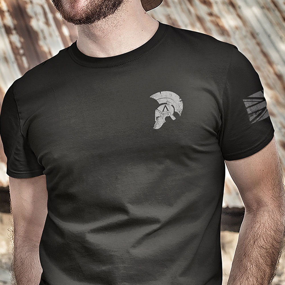 Close up of Front of Man wearing Black short sleeve unisex fit cotton T-Shirt by Achilles Tactical Clothing Brand printed with Achilles Helmet logo design across the front left chest